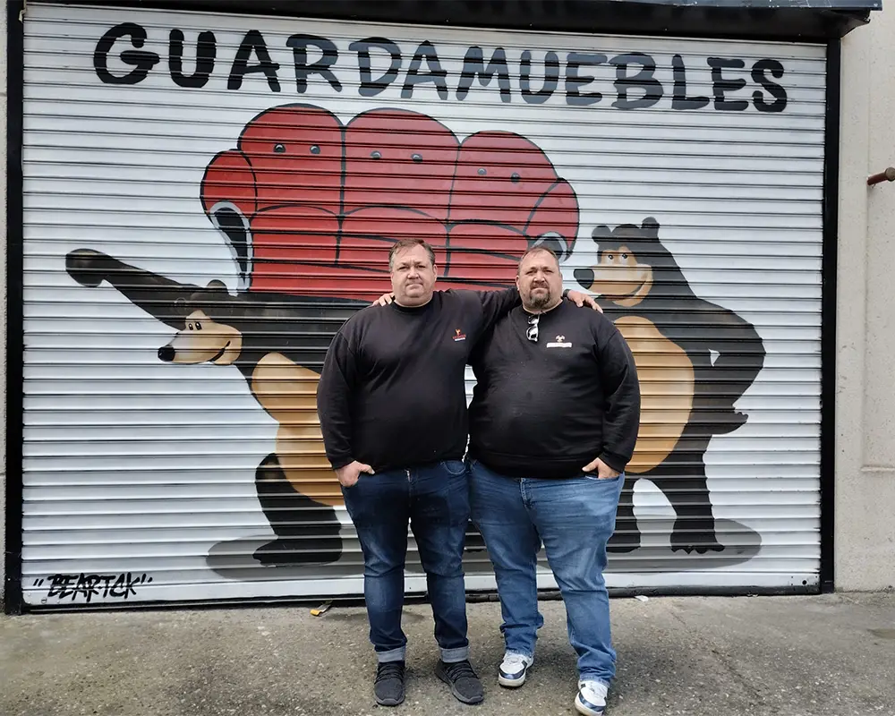 Los Gemelos frente a una persiana metálica con un mural pintado que dice 'GUARDAMUEBLES'