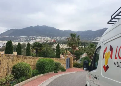 Furgoneta de mudanzas de Los Gemelos aparcada frente a una residencia con montañas brumosas en el fondo.